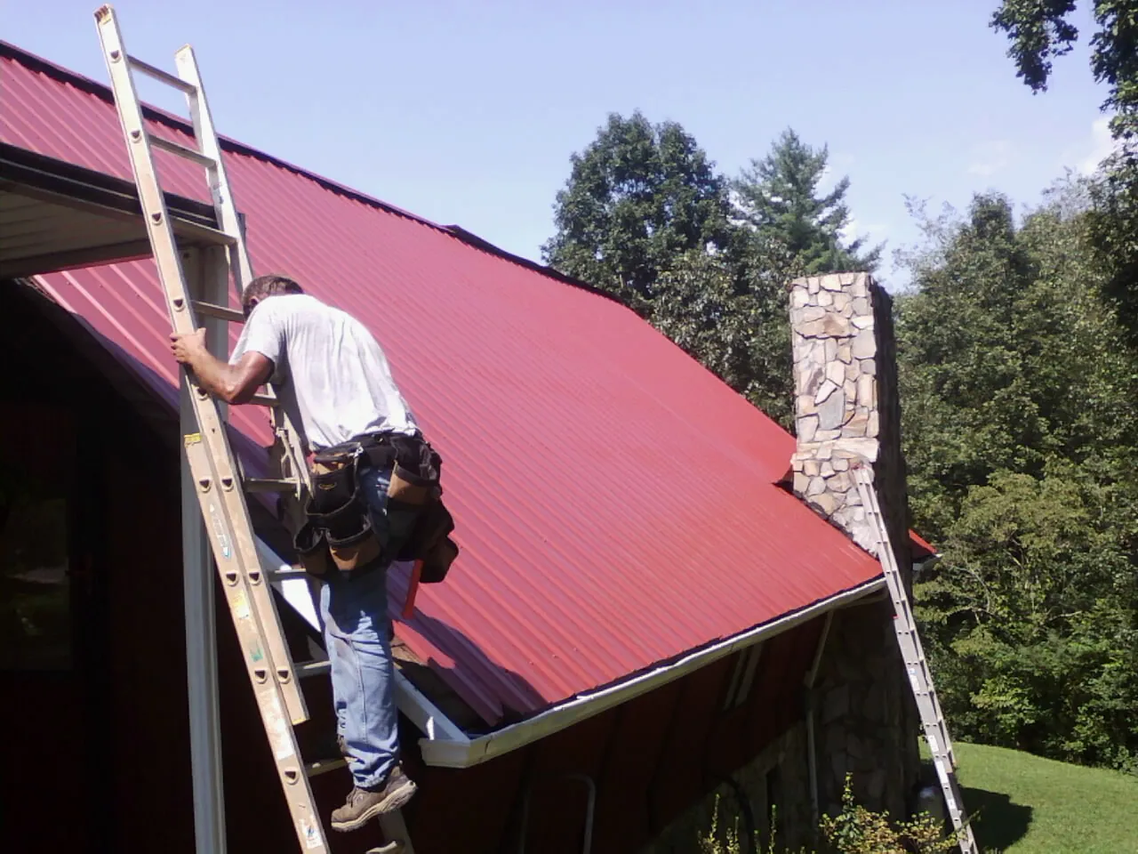 Roofer on a ladder beside a red metal roof and stone chimney — Soffit & Fascia Repair by Roofing Brothers in Saint Paul Park